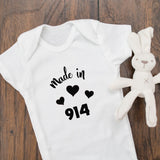 A white baby bodysuit laid flat on a wooden surface, printed with the words 'Made in 914' and small black hearts. A small white stuffed bunny toy sits to the right of the bodysuit.
