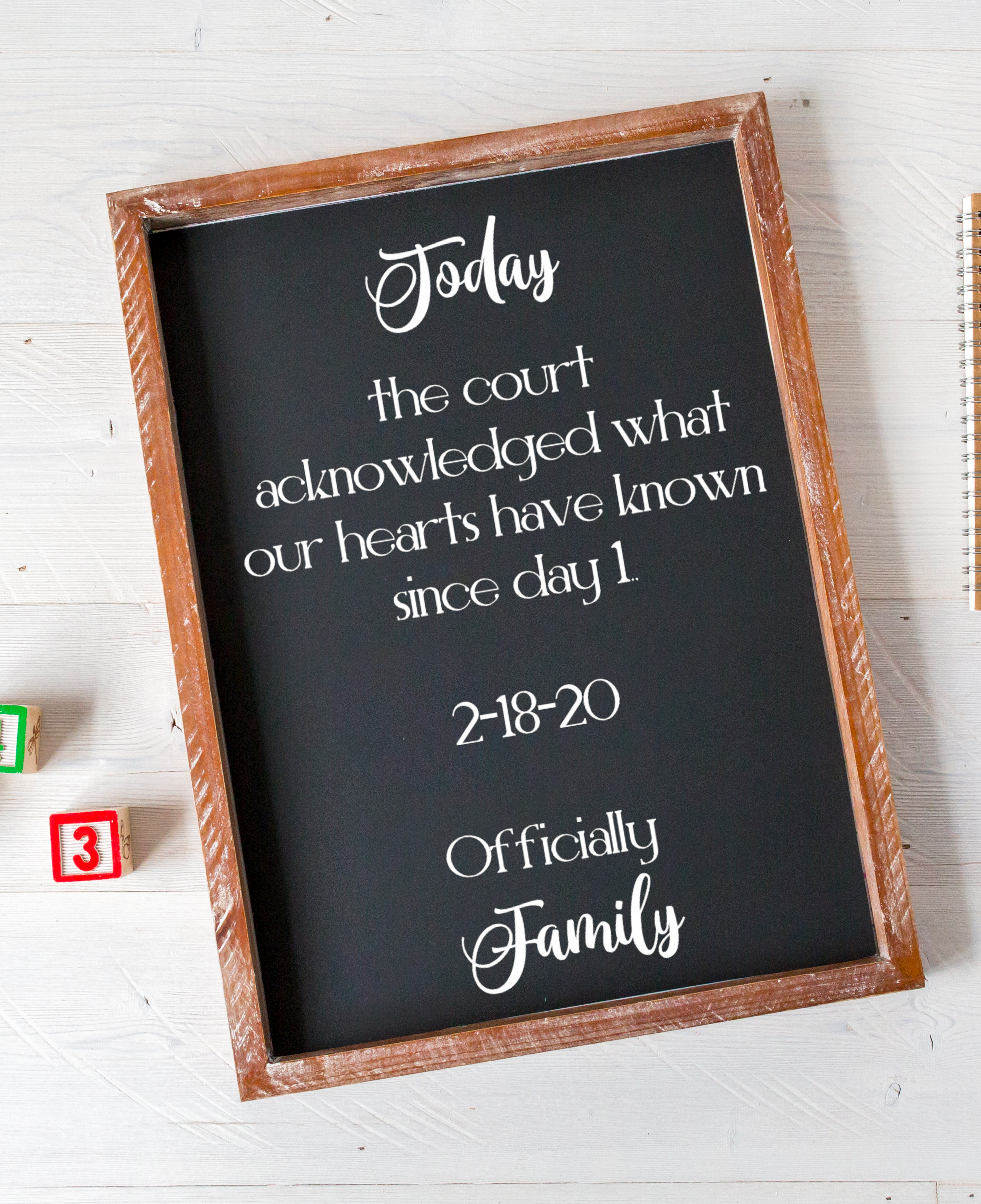 A rectangular chalkboard-style sign with a rustic wooden frame, printed with white script and block text reading 'Today the court acknowledged what our hearts have known since day 1 2-18-20 Officially Family.' It is photographed on a white painted wood surface with small colorful blocks partly visible at the lower left.