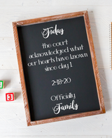A rectangular chalkboard-style sign with a rustic wooden frame, printed with white script and block text reading 'Today the court acknowledged what our hearts have known since day 1 2-18-20 Officially Family.' It is photographed on a white painted wood surface with small colorful blocks partly visible at the lower left.