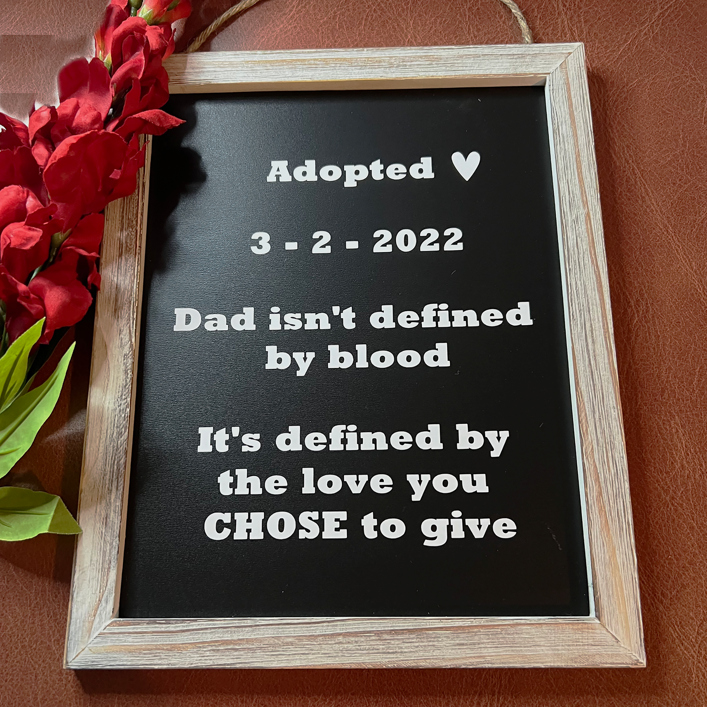A framed black sign with white text reading 'Adopted 3-2-2022 Dad isn't defined by blood It's defined by the love you CHOSE to give', shown in a light wood frame on a brown textured background with red flowers at the side.