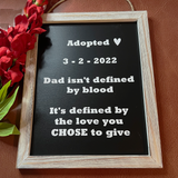 A framed black sign with white text reading 'Adopted 3-2-2022 Dad isn't defined by blood It's defined by the love you CHOSE to give', shown in a light wood frame on a brown textured background with red flowers at the side.