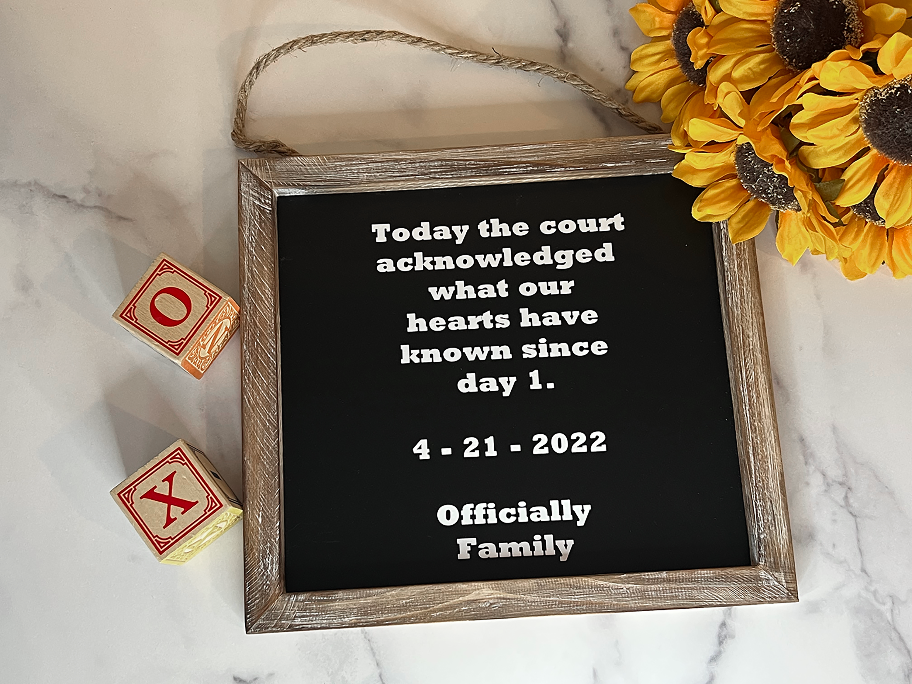A rustic wooden framed black sign with white text reading a court acknowledgment date and phrase, styled on a white marble surface with sunflowers and wooden blocks.