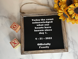 A rustic wooden framed black sign with white text reading a court acknowledgment date and phrase, styled on a white marble surface with sunflowers and wooden blocks.