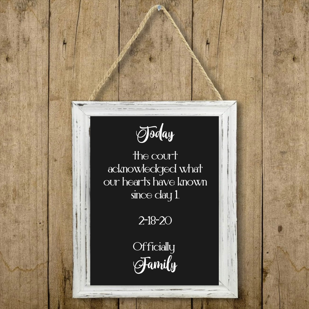 A framed hanging sign with a black chalkboard-style center and white script text reading a family affirmation, shown against a rustic wood plank background. The sign has a distressed white frame and hangs from a rope.