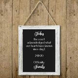 A framed hanging sign with a black chalkboard-style center and white script text reading a family affirmation, shown against a rustic wood plank background. The sign has a distressed white frame and hangs from a rope.