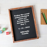 A framed black chalkboard-style sign with white text reading 'Today the court acknowledged what our hearts have known since day 1. 4 - 21 - 2022 Officially Family,' photographed on a white wood surface with small toy blocks and a notebook with pencils nearby.
