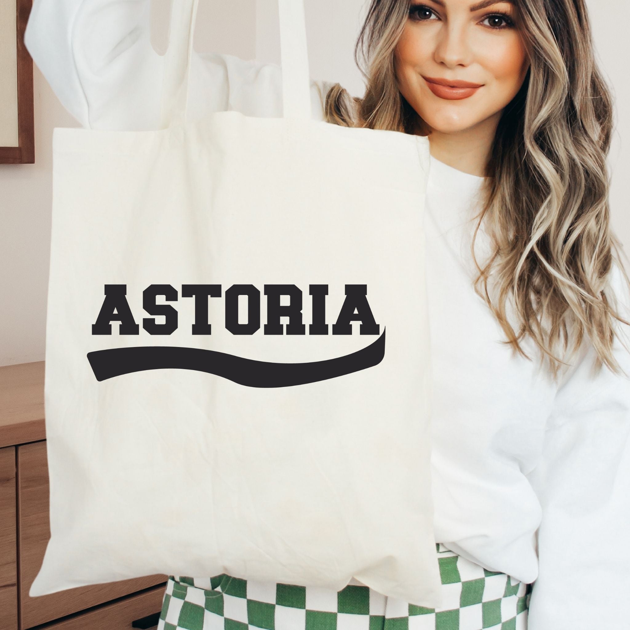 A woman holding a cream canvas tote bag with the word 'ASTORIA' printed in bold black letters across the front, wearing a white sweatshirt and green-and-white checkered pants.