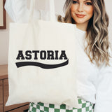 A woman holding a cream canvas tote bag with the word 'ASTORIA' printed in bold black letters across the front, wearing a white sweatshirt and green-and-white checkered pants.