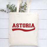 An off-white canvas tote bag with the word 'ASTORIA' printed in red across the front, hanging against a white wood plank background with green eucalyptus stems at the top.