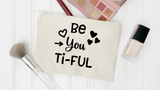 A cream canvas makeup bag with black text reading 'Be You Ti-FUL' surrounded by heart and arrow graphics, styled in a flat-lay with makeup accessories on a white wood background.