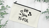 Canvas Natural pouch with black wriitng Be You Ti-Ful and hearts around it, with a white wood background and green leaves on the corners.