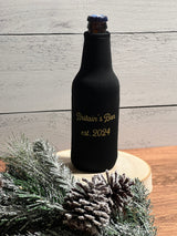 A black neoprene bottle sleeve with gold lettering reading 'Britain's Bar est. 2024' on a brown glass bottle, styled on a wooden slice with pinecones and frosted greenery against a light wood plank background.