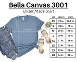 A unisex fit size chart graphic for Bella Canvas 3001, showing a blue T-shirt with measurement lines and a size table listing XS through 4X with length and width measurements. Jeans, sandals, and a small plant are styled around the shirt on a white background.