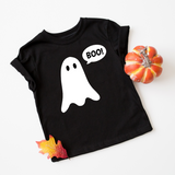 A black short-sleeve baby t-shirt with a white ghost graphic and a speech bubble that says 'BOO!' in all caps. The t-shirt is styled in a flat-lay on a white background, decorated with a small orange pumpkin and an autumn leaf.