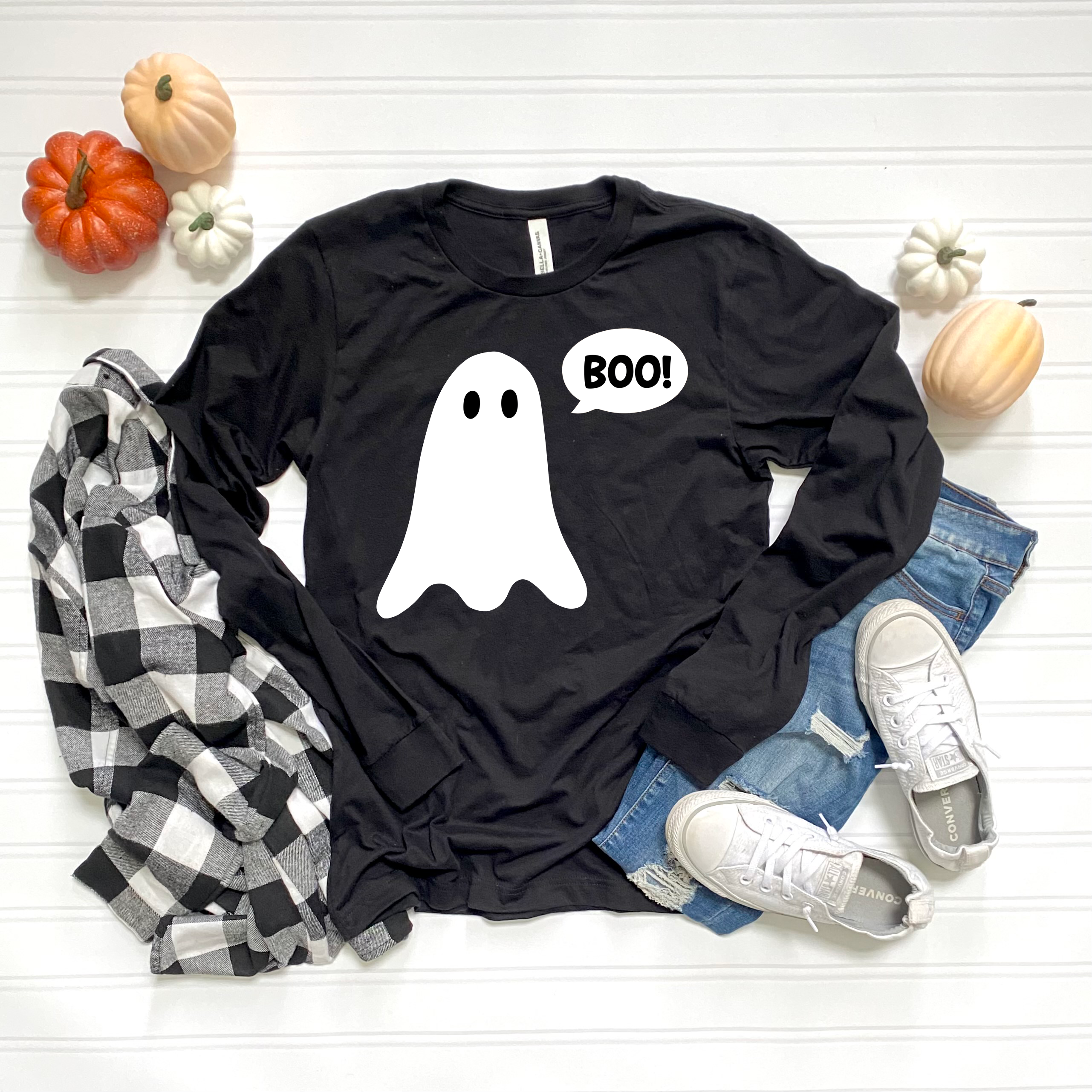 A black long-sleeve graphic T-shirt with a white ghost and the word 'BOO!' in a speech bubble, styled in a flat lay with a black-and-white plaid shirt, blue jeans, white sneakers, and small pumpkins on a white wood background.