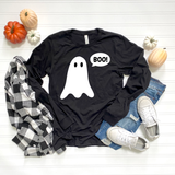 A black long-sleeve graphic T-shirt with a white ghost and the word 'BOO!' in a speech bubble, styled in a flat lay with a black-and-white plaid shirt, blue jeans, white sneakers, and small pumpkins on a white wood background.