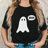 A woman wearing a black short-sleeve t-shirt with a white cartoon ghost graphic and a speech bubble that says 'BOO!'. She is wearing blue jeans and standing in front of a light wood paneled wall.