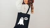 A woman wearing a white ribbed sweater and a brown hat carries a black canvas tote bag with a white ghost graphic and a speech bubble that says 'BOO!'.
