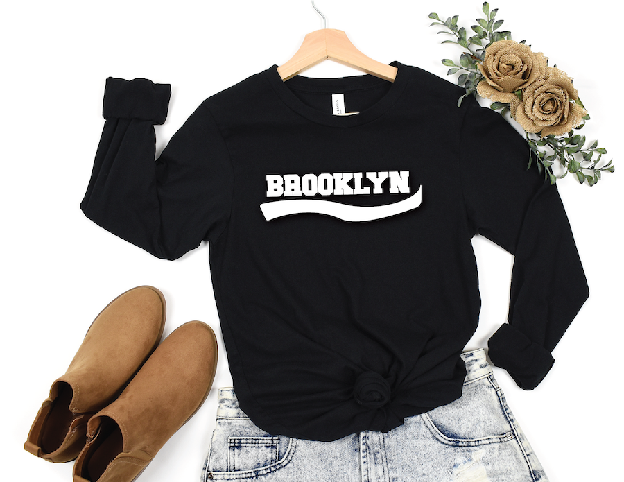A black long-sleeve crewneck sweatshirt with 'BROOKLYN' printed in white across the chest, styled in a flat lay on a wooden hanger with light-wash jeans, tan suede ankle boots, and dried flowers on a white background.