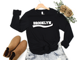 A black long-sleeve crewneck sweatshirt with 'BROOKLYN' printed in white across the chest, styled in a flat lay on a wooden hanger with light-wash jeans, tan suede ankle boots, and dried flowers on a white background.