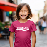 A young girl with brown hair smiling in a blurred outdoor street setting, wearing a bright pink t-shirt with 'BROOKLYN' printed in white across the chest.