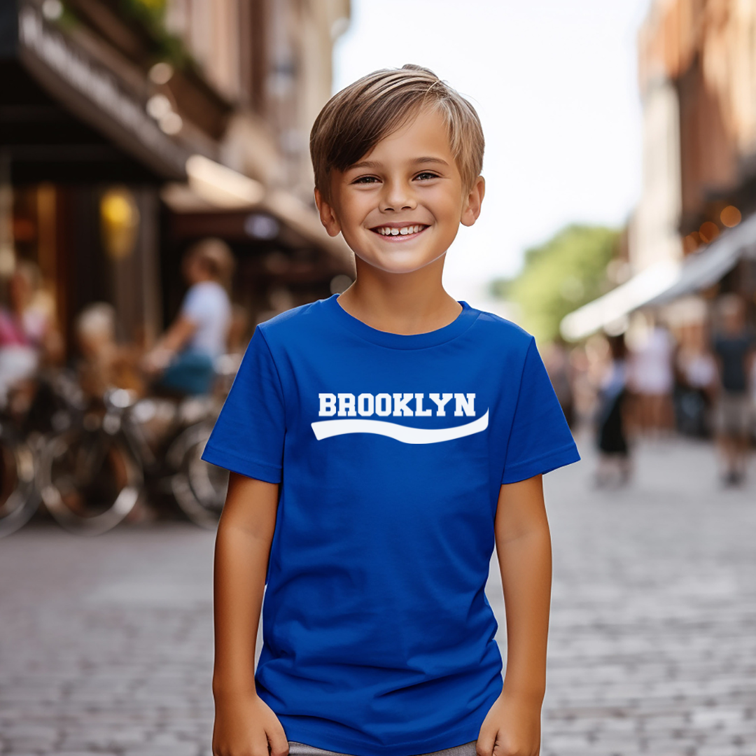 A young boy smiling in a cobblestone street wearing a royal blue t-shirt with 'BROOKLYN' printed in white across the chest.