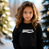 A young girl with long, wavy light brown hair is wearing a black crewneck sweatshirt with 'BROOKLYN' printed in white across the chest. The background is a soft-focus outdoor winter scene with snow and decorated Christmas trees.