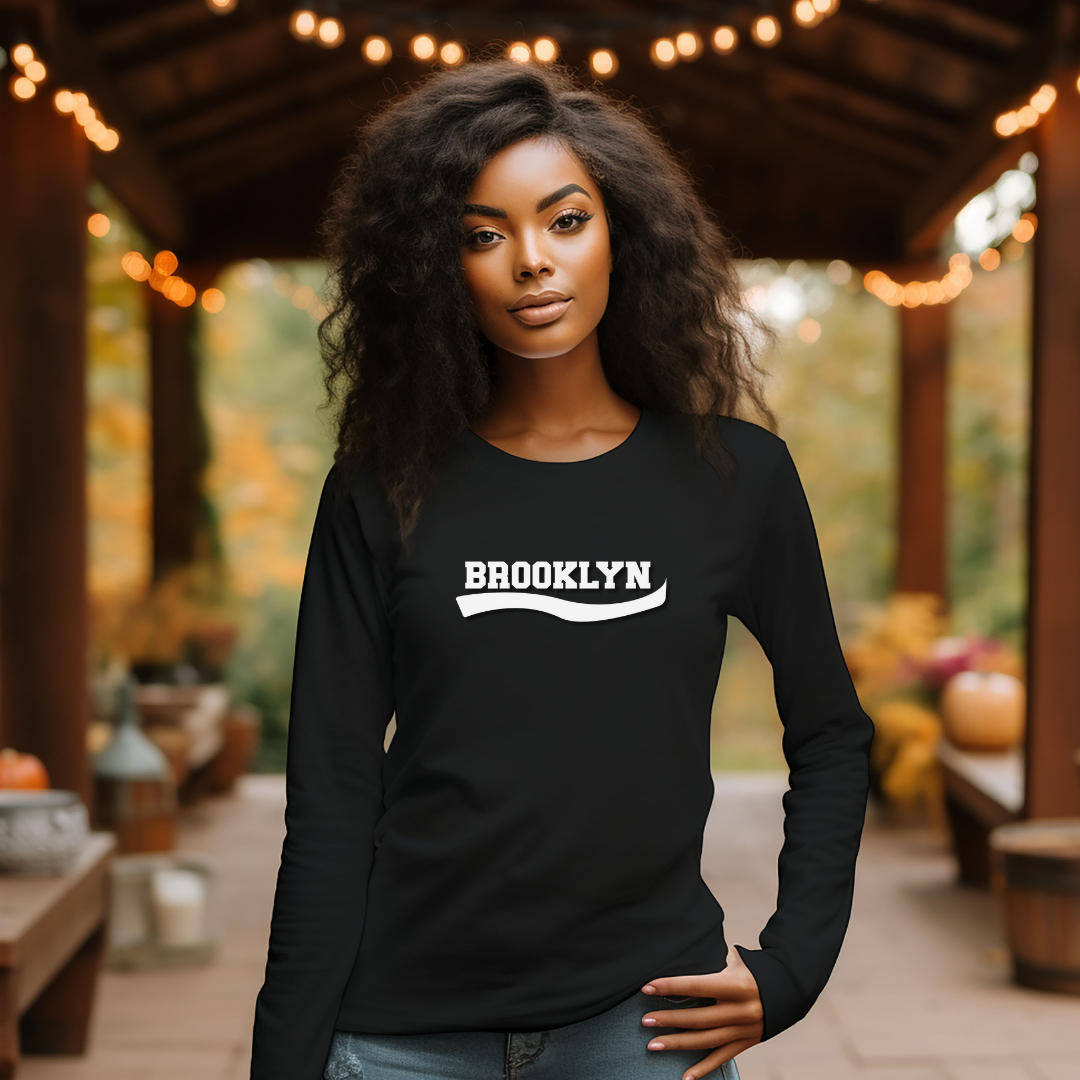 A woman wearing a black long-sleeve crewneck sweatshirt with 'BROOKLYN' printed in white across the chest, standing on a wooden covered patio with warm string lights and autumn foliage in the background.