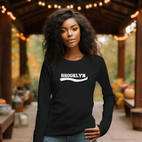 A woman wearing a black long-sleeve crewneck sweatshirt with 'BROOKLYN' printed in white across the chest, standing on a wooden covered patio with warm string lights and autumn foliage in the background.