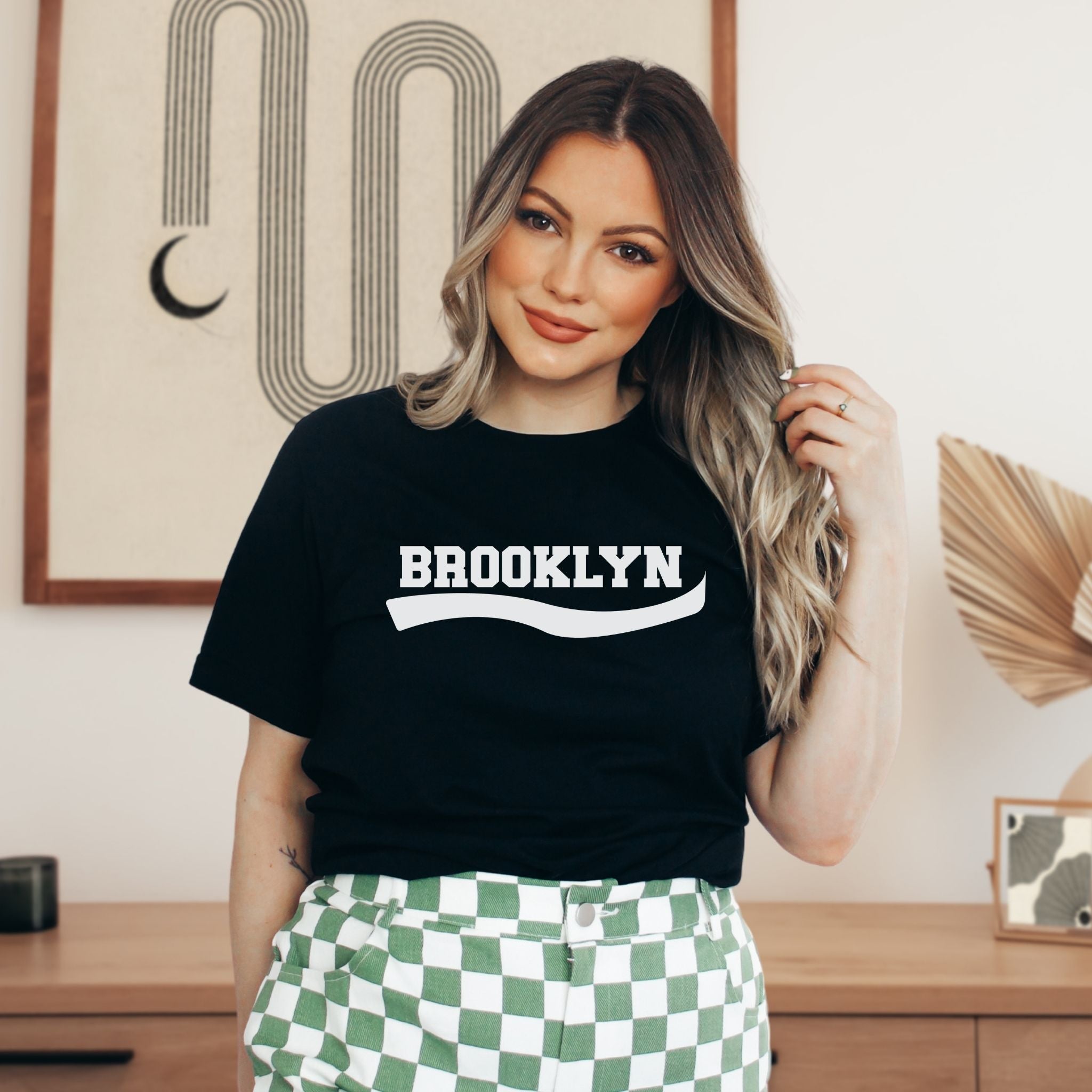 A woman wearing a black crewneck t-shirt with 'BROOKLYN' printed in white across the chest, paired with green and white checkered pants, standing in a styled indoor room.