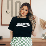 A woman wearing a black crewneck t-shirt with 'BROOKLYN' printed in white across the chest, paired with green and white checkered pants, standing in a styled indoor room.