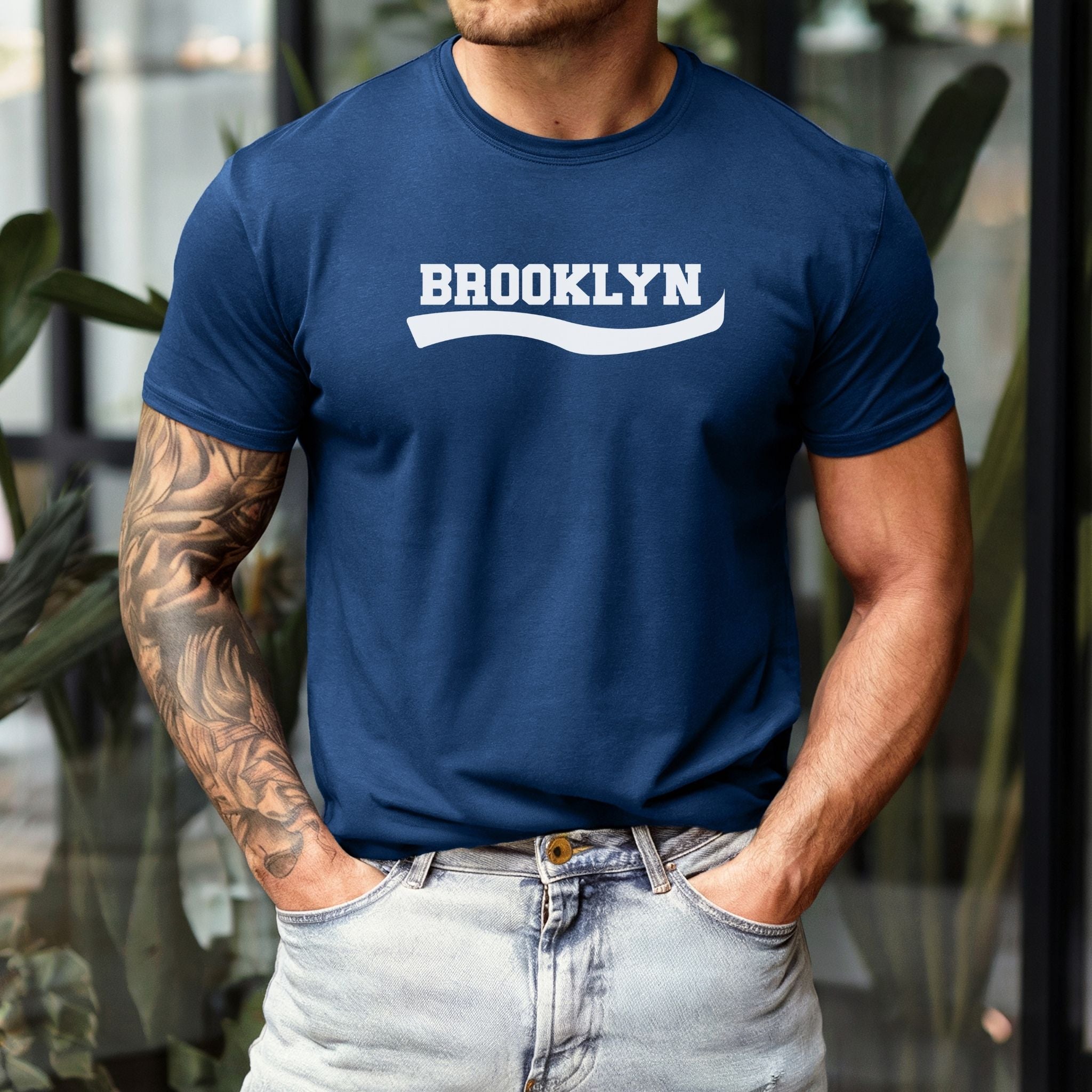 A man wearing a navy blue crewneck t-shirt with 'BROOKLYN' printed in white across the chest, paired with light-wash jeans.