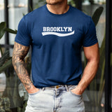 A man wearing a navy blue crewneck t-shirt with 'BROOKLYN' printed in white across the chest, paired with light-wash jeans.