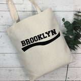 An off-white canvas tote bag with 'BROOKLYN' printed in black across the front, resting on a white wood plank surface next to a green eucalyptus sprig.