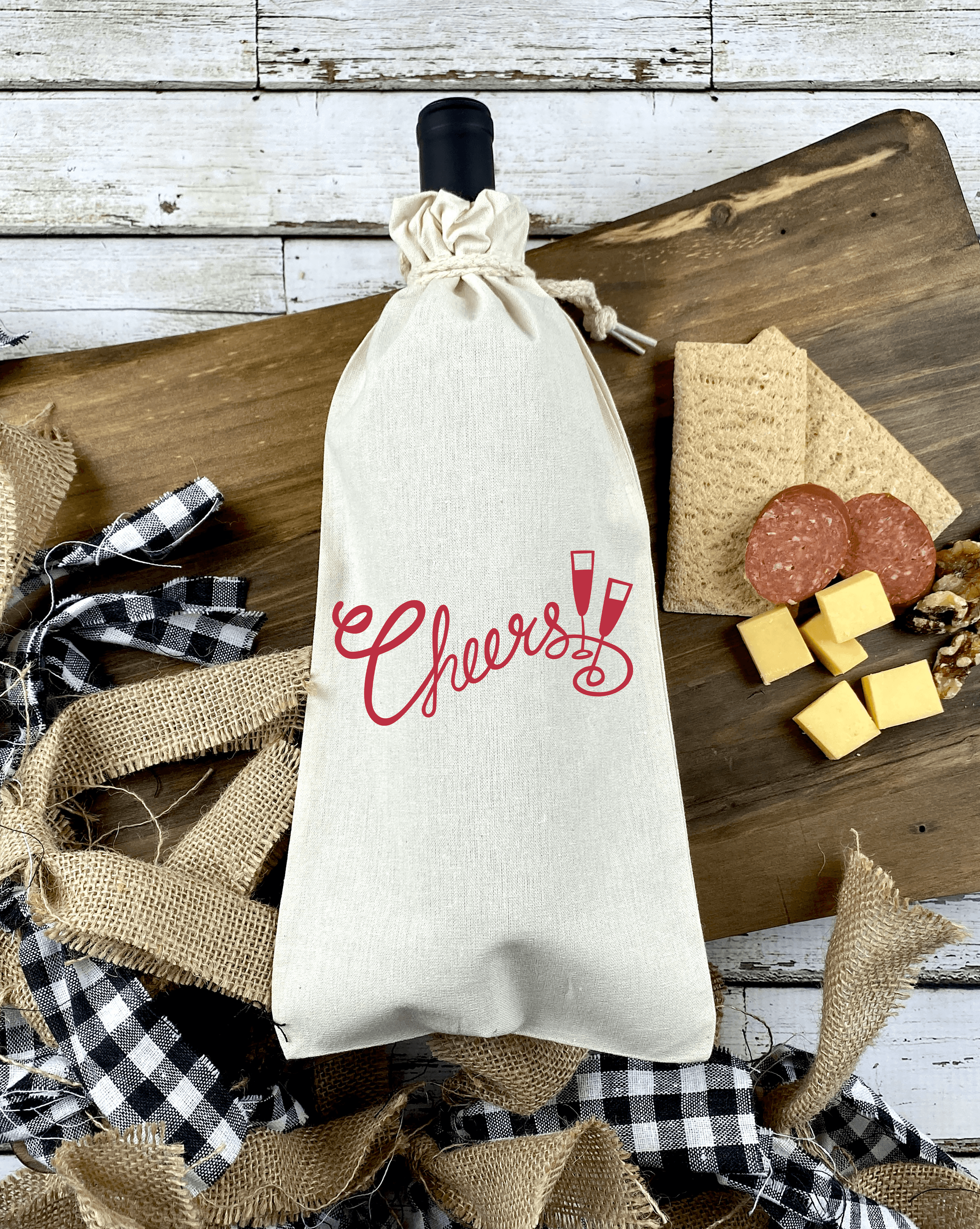 Cloth wine bag with red words Cheers and two glasses in the middle with wine bottle inside, resting over a chatturrie board in wood and chees, nuts, salami and crakcers to the right and buffalo plaid ribbon on the left.