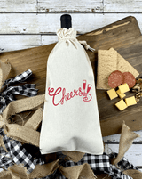 Cloth wine bag with red words Cheers and two glasses in the middle with wine bottle inside, resting over a chatturrie board in wood and chees, nuts, salami and crakcers to the right and buffalo plaid ribbon on the left.