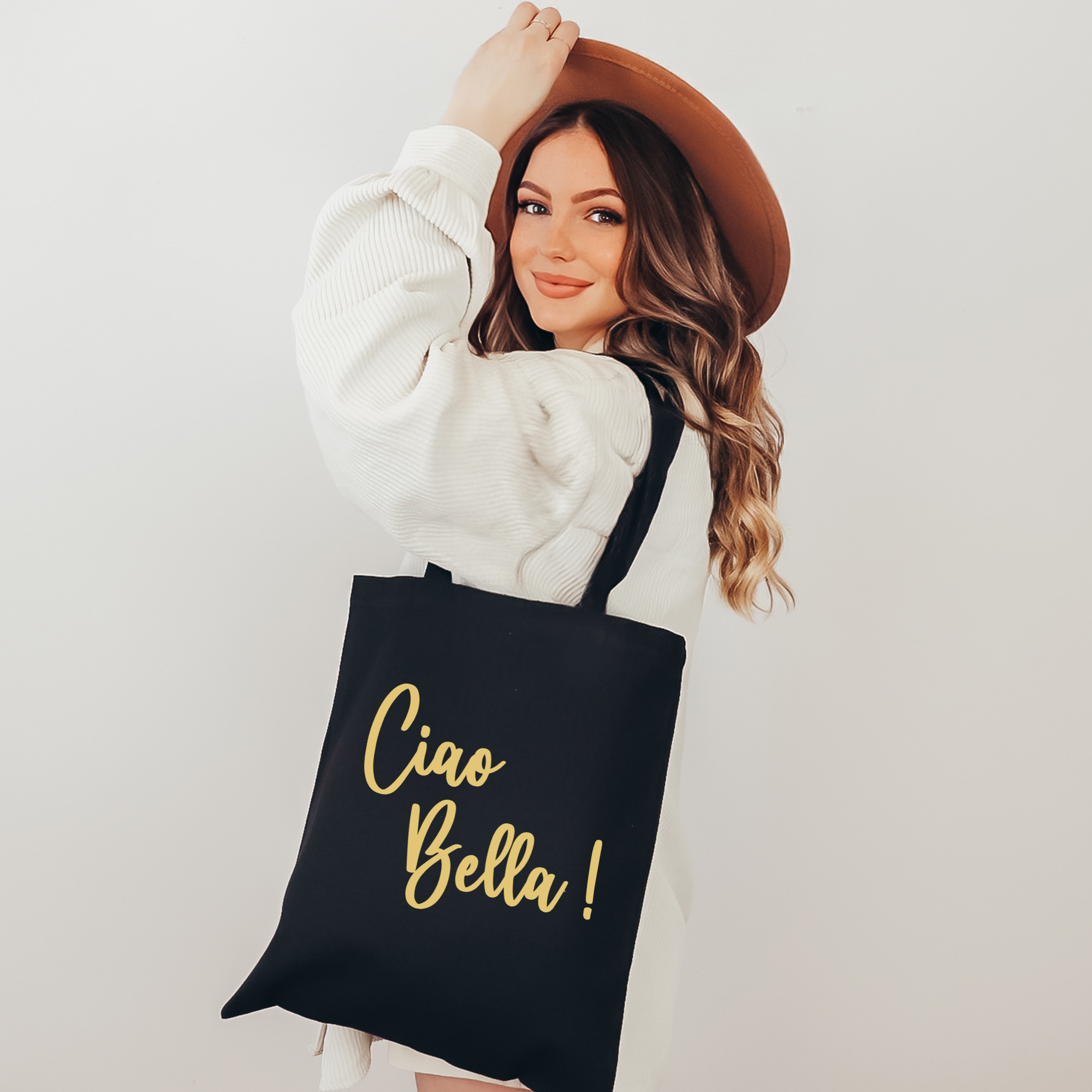 A woman wearing a brown wide-brim hat and a white ribbed sweater holds a black canvas tote bag with gold foil text that reads 'Ciao Bella!'.