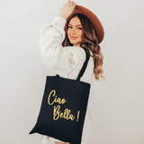 A woman wearing a brown wide-brim hat and a white ribbed sweater holds a black canvas tote bag with gold foil text that reads 'Ciao Bella!'.