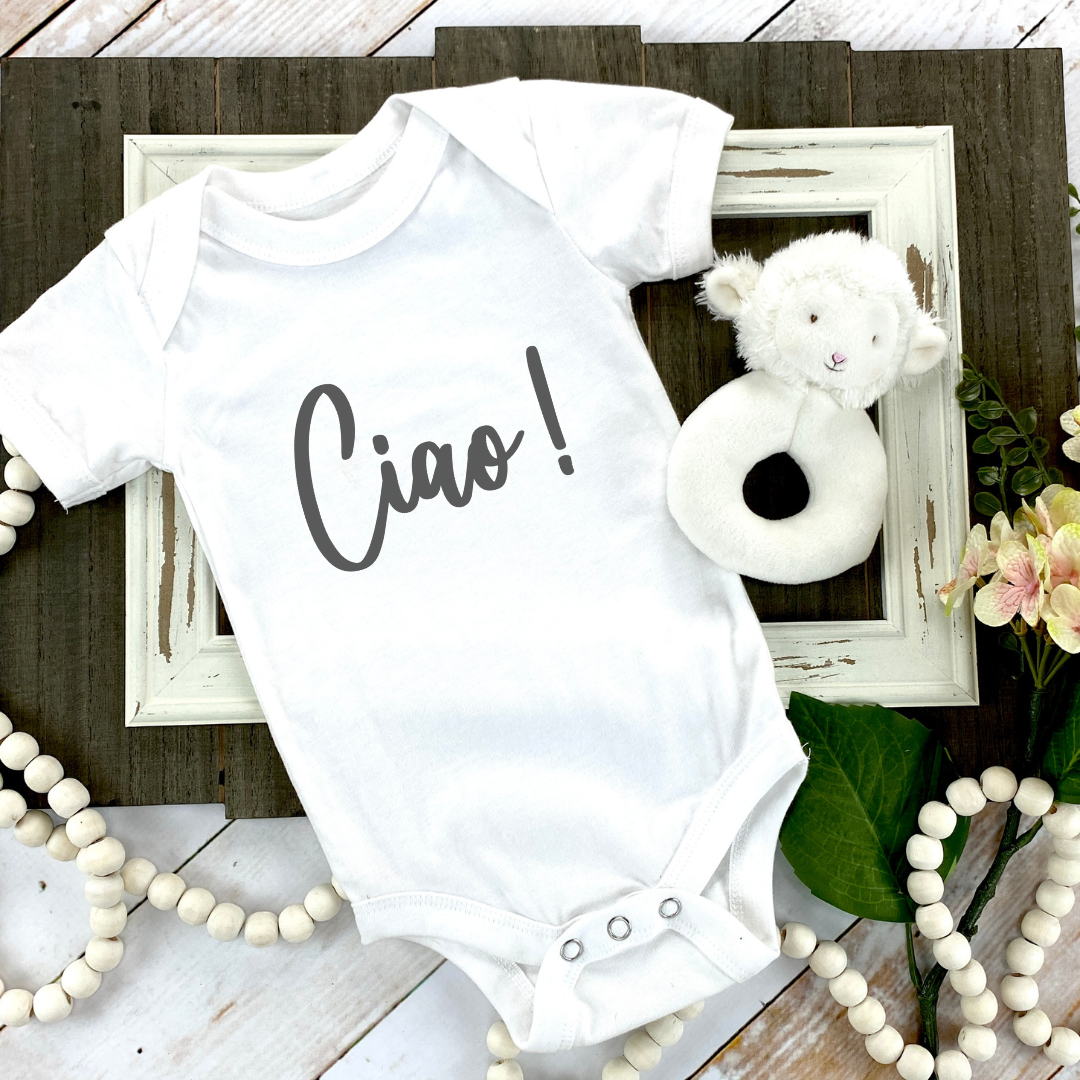 A white short-sleeve baby bodysuit laid flat on a rustic wood surface, printed with the word 'Ciao!' in dark gray script. The bodysuit is styled with a distressed white picture frame, a small plush sheep toy, green leaves, pink flowers, and white bead garlands.
