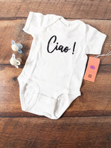 A white short-sleeve baby bodysuit laid flat on a wooden surface, printed with the word 'Ciao!' in black script. Two pacifiers and a peach-colored product tag are placed beside it.