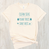 A cream-colored short-sleeve t-shirt with a teal graphic print that reads 'CLEAN SEAS PLANT TREES SAVE BEES' with simple illustrations of trees, bees, and a wave, laid flat on a light wood background.