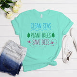A mint green graphic t-shirt laid flat on a white wood background, styled with a pair of blue denim shorts, white sneakers, and yellow roses. The t-shirt features the text 'CLEAN SEAS PLANT TREES SAVE BEES' in blue, green, and pink lettering.