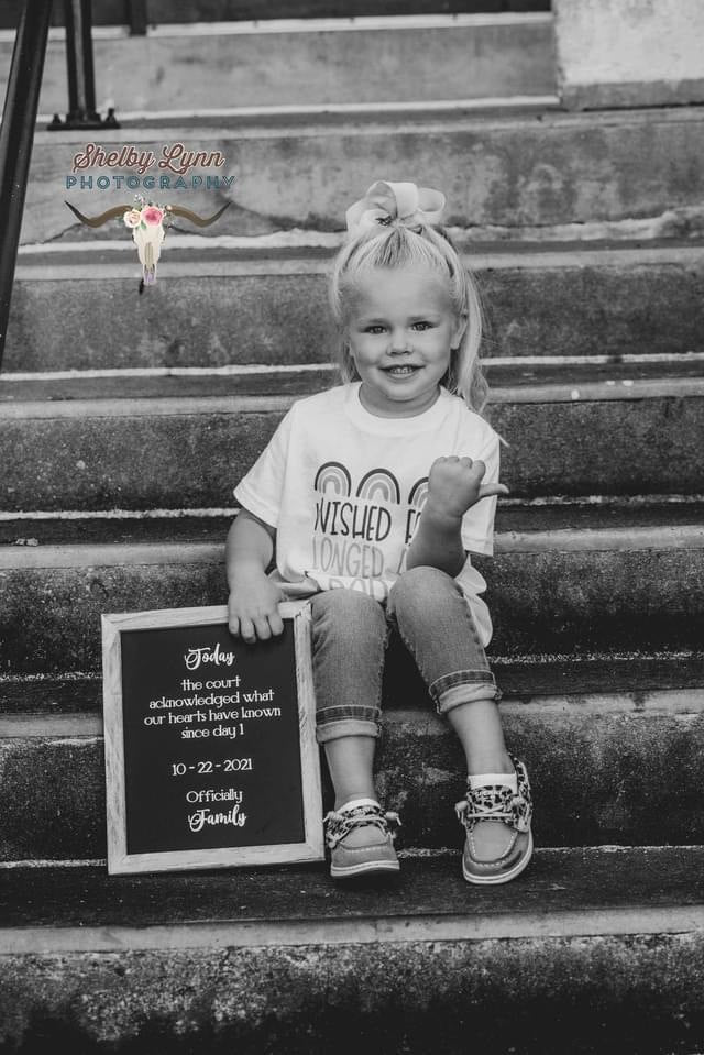 A black and white photograph of a young girl sitting on outdoor stone steps, holding a small framed sign that reads 'Today the court declared our hearts have tattoos since day 1 10-29-2019 Officially Family'. She is wearing a white graphic t-shirt, rolled-up jeans, and sneakers, with a large hair bow.