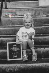 A black and white photograph of a young girl sitting on outdoor stone steps, holding a small framed sign that reads 'Today the court declared our hearts have tattoos since day 1 10-29-2019 Officially Family'. She is wearing a white graphic t-shirt, rolled-up jeans, and sneakers, with a large hair bow.