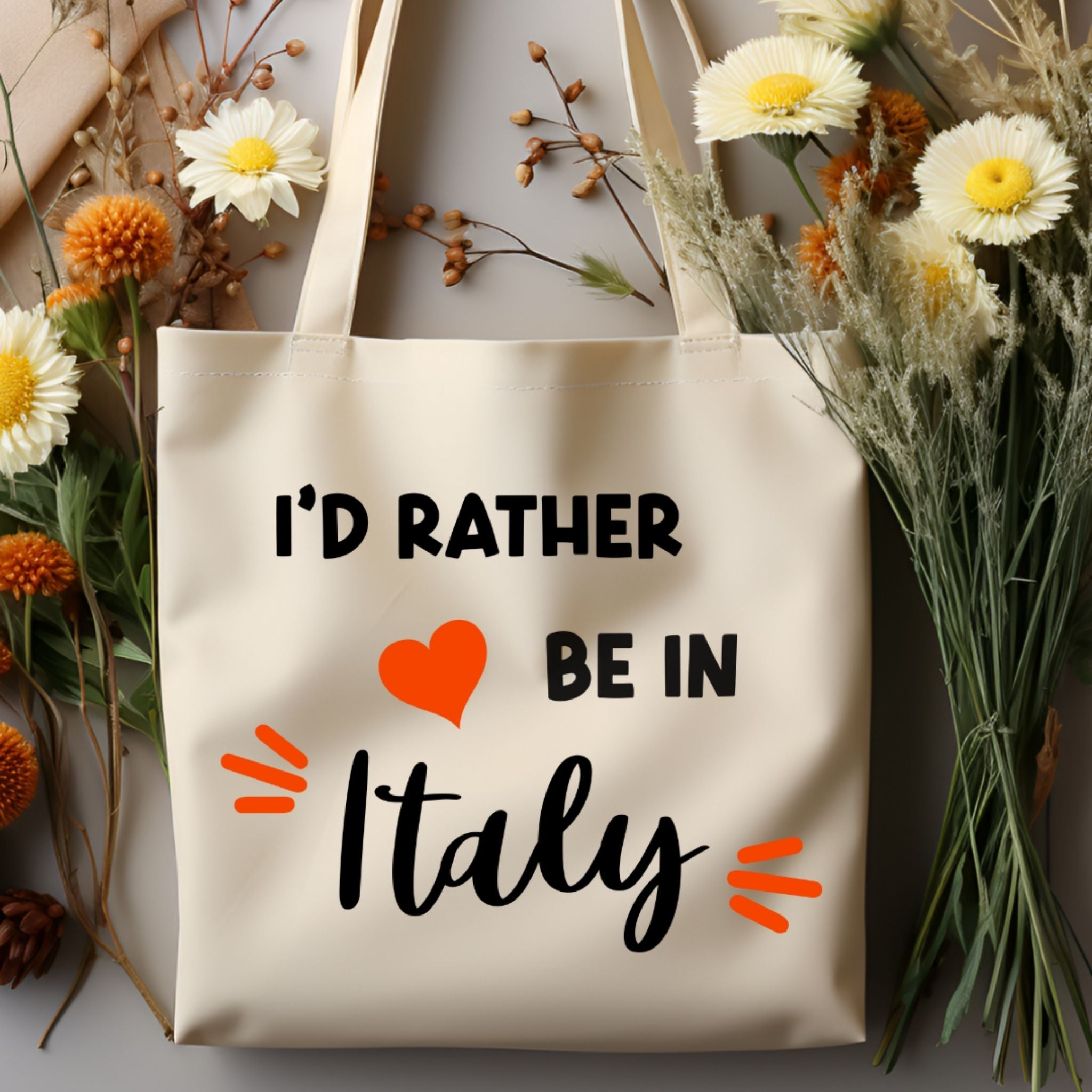 A cream canvas tote bag with the text 'I'D RATHER BE IN Italy' in black, featuring a red heart and orange accent marks. The bag is styled in a flat-lay arrangement with white and yellow daisies and dried flowers on a gray surface.