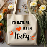 A cream canvas tote bag with the text 'I'D RATHER BE IN Italy' in black, featuring a red heart and orange accent marks. The bag is styled in a flat-lay arrangement with white and yellow daisies and dried flowers on a gray surface.