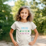A young girl with wavy brown hair stands outdoors wearing a cream-colored t-shirt with a colorful graphic that reads 'CLEAN SEAS PLANT TREES SAVE BEES' in blue, green, and pink text.
