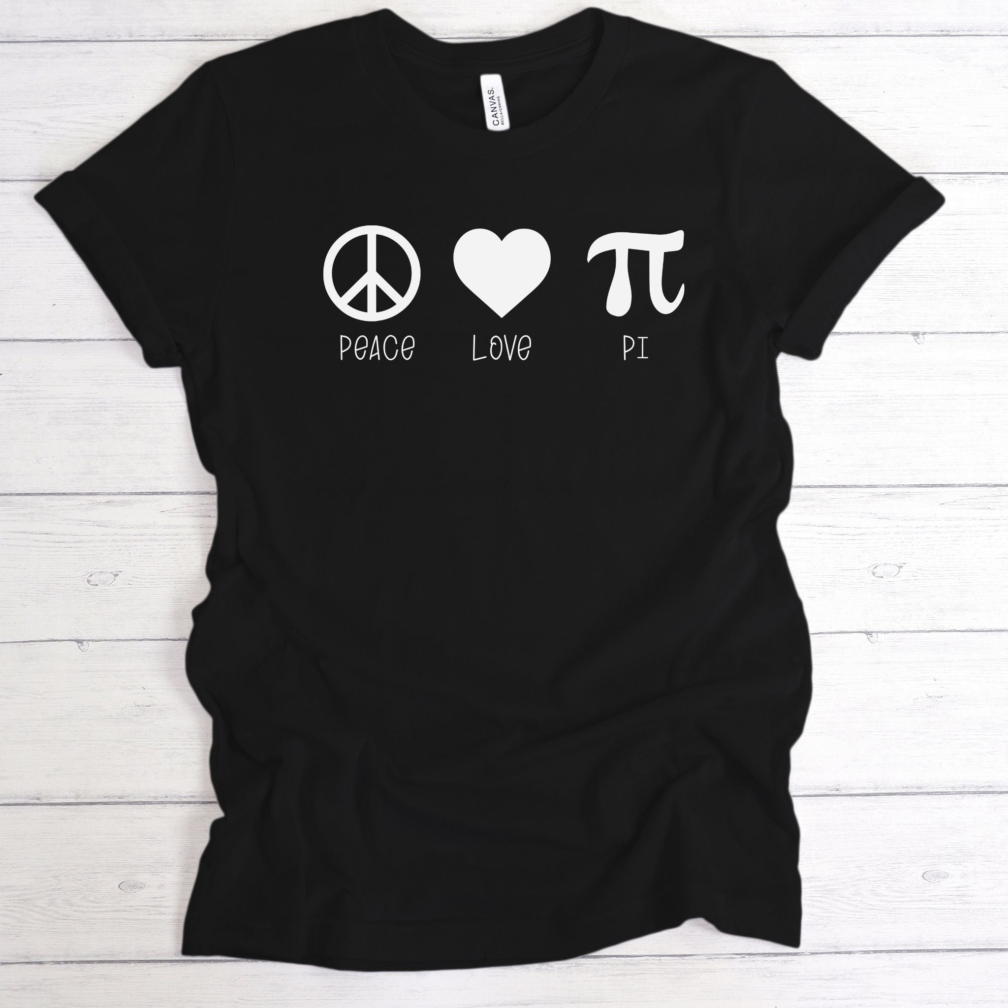 A black short-sleeve t-shirt laid flat on a white wood plank background, featuring a white graphic print of a peace symbol, a heart, and a pi symbol with the words 'PEACE', 'LOVE', and 'PI' underneath.