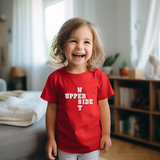 A young child with light brown hair is smiling and laughing while wearing a red t-shirt with white text that reads 'UPPER SIDE T'. The background shows a softly blurred, warm-toned home interior with a bookshelf, curtains, and a bed.