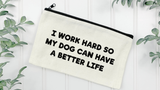 An off-white canvas zipper pouch with black text reading 'I WORK HARD SO MY DOG CAN HAVE A BETTER LIFE' on a white paneled background, surrounded by green leaves.